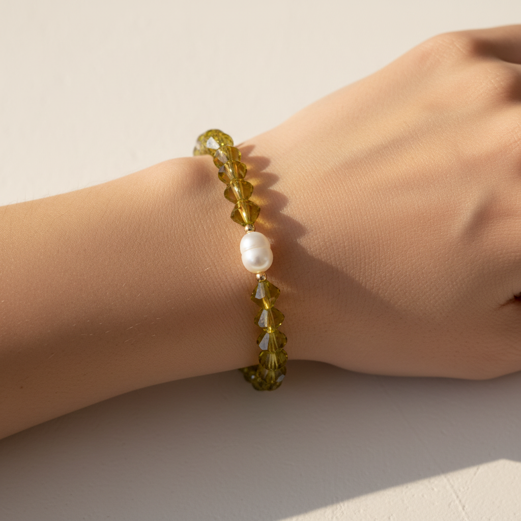 The Juniper Bracelet on Wrist. This is an arm sitting on a white background with a shadow. The beads on the wrist are green in an unique shape. A fresh water pearl is front in center surrounded by gold filled beads.