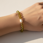 The Juniper Bracelet on Wrist. This is an arm sitting on a white background with a shadow. The beads on the wrist are green in an unique shape. A fresh water pearl is front in center surrounded by gold filled beads.