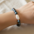 The Lyra Bracelet on wrist. This is a close up of the bracelet. it is sitting on the wrist with an ivory fabirc as the background. The bracelet is an unique shade of blues, greens and purples swirled together with a long rectangle. Gold sapcers surrounding the pearl.