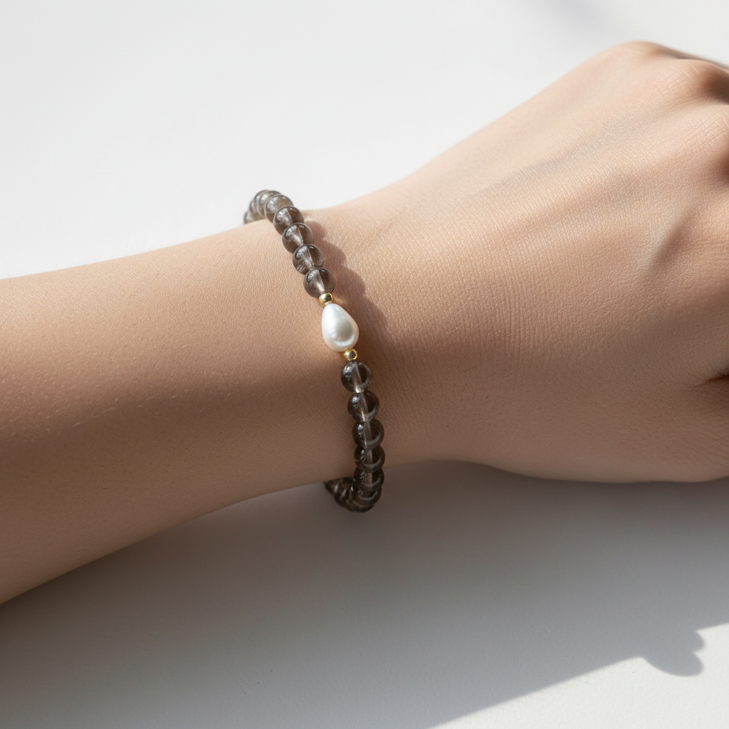 A close up of a feminine wrist sitting on white table. Bracelet on wrist is smoky brown with a fresh water pearl teardrop shape. Gold filled beads surround the pearl. 
