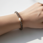 A close up of a feminine wrist sitting on white table. Bracelet on wrist is smoky brown with a fresh water pearl teardrop shape. Gold filled beads surround the pearl. 