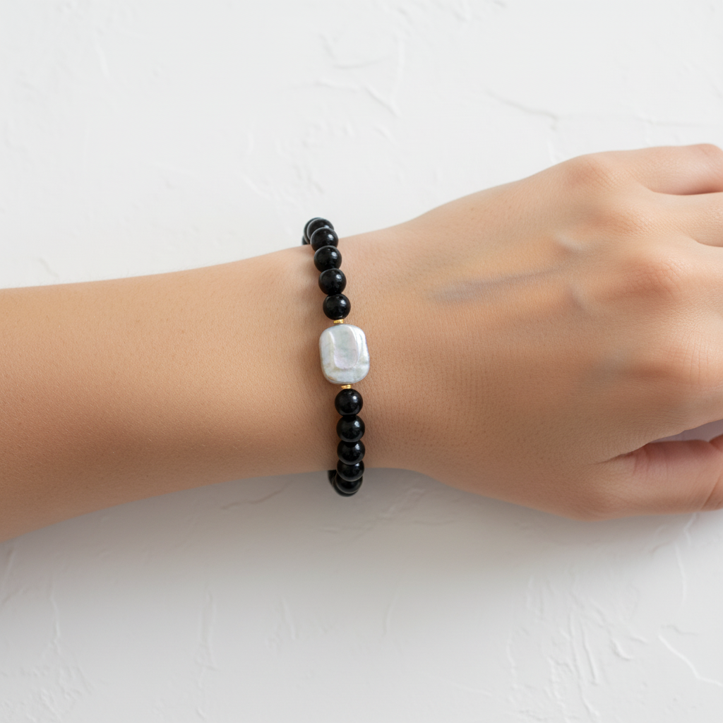 The Vesper Bracelet - wrist shot. On a female wrist is black shiny beads and a large rectanle resh water pearl. Gold spacers surrounding pearl.