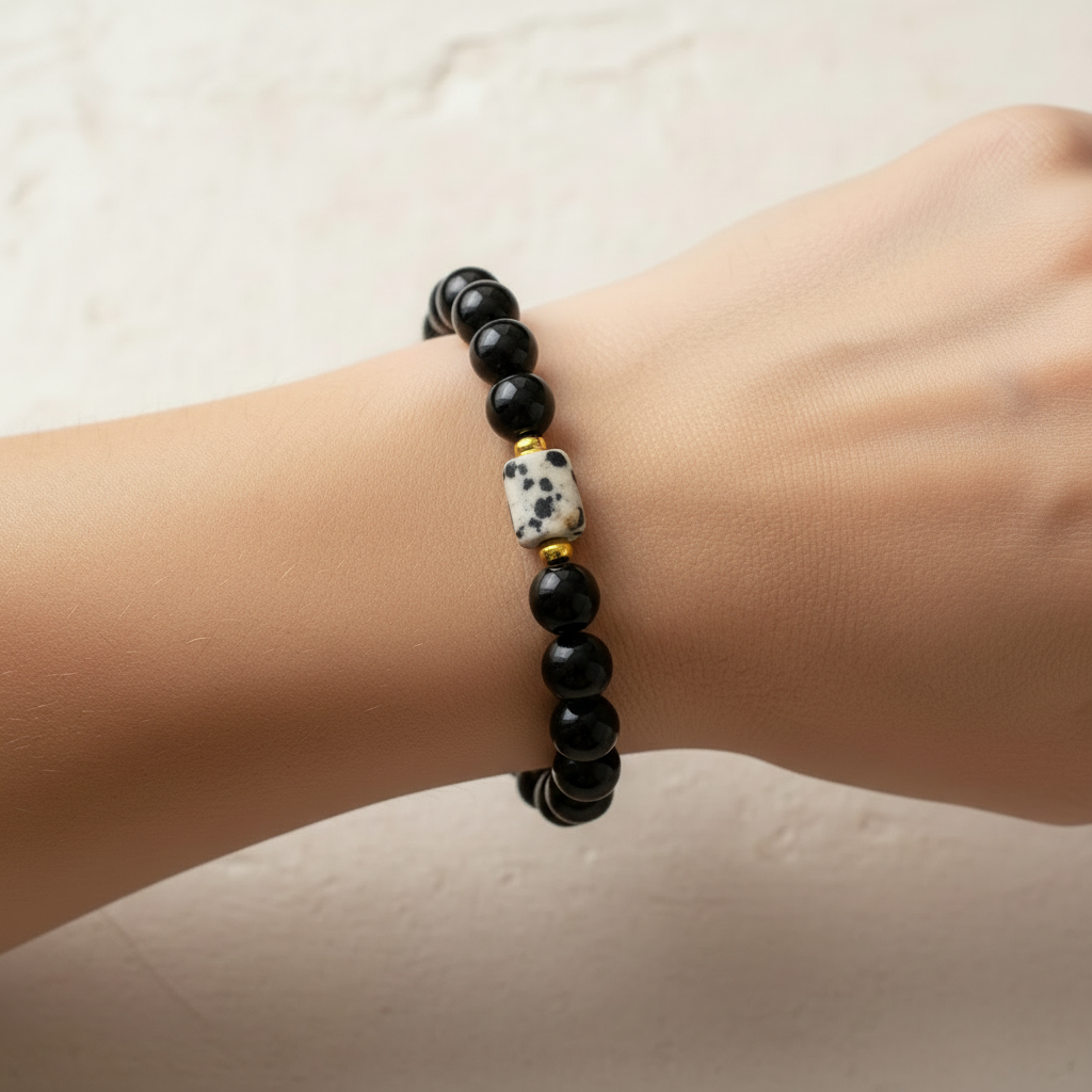 The Ziva Bracelet on wrist - gold spacers. This is a close up of a wrist. The bracelet on the wrist is black with a small rectangle spotted bead. Gold spacers surroundthe accent.